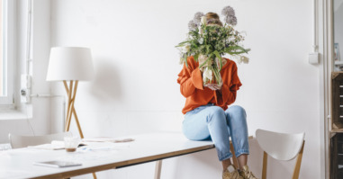 Young woman sitting on table holding flower vase in front of her face