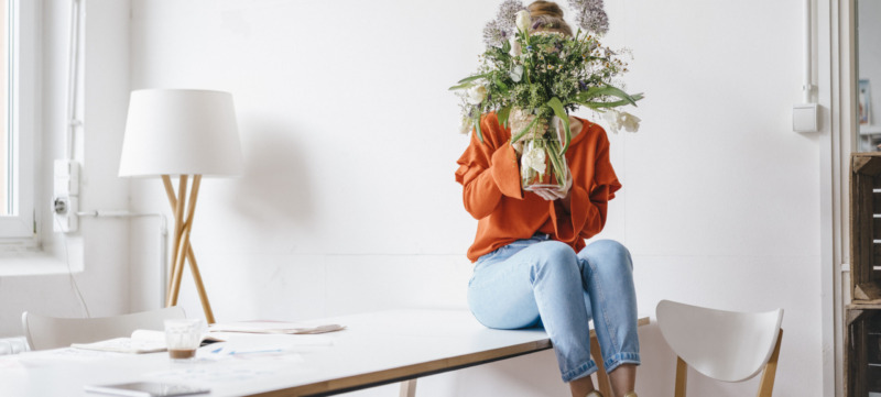 Young woman sitting on table holding flower vase in front of her face