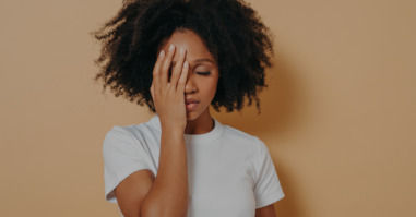 Dark skinned female covering face with hand and feeling anxiety or depression against beige wall