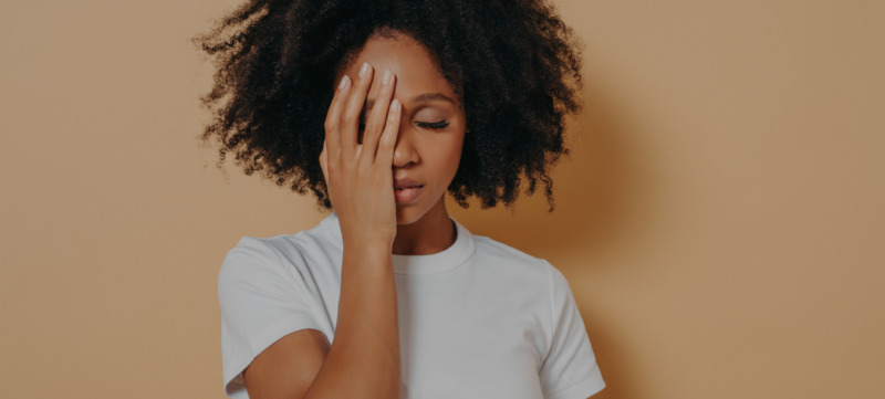 Dark skinned female covering face with hand and feeling anxiety or depression against beige wall