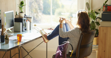 Pensive woman looking through window with feet up on desk in sunny home office