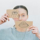 Vrouw heeft voor haar oog en mond een papiertje. Met daarop een getekend oog en getekende mond. Spelend met de verhoudingen in haar gezicht.