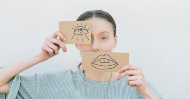 Vrouw heeft voor haar oog en mond een papiertje. Met daarop een getekend oog en getekende mond. Spelend met de verhoudingen in haar gezicht.