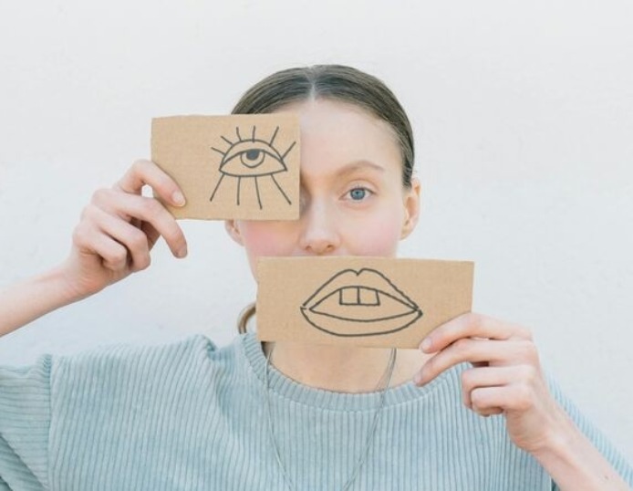 Vrouw heeft voor haar oog en mond een papiertje. Met daarop een getekend oog en getekende mond. Spelend met de verhoudingen in haar gezicht.