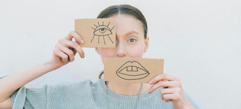 Vrouw heeft voor haar oog en mond een papiertje. Met daarop een getekend oog en getekende mond. Spelend met de verhoudingen in haar gezicht.